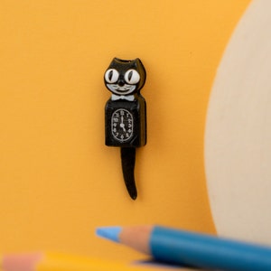 Può includere: Un orologio nero a forma di gatto con papillon bianco e grandi occhi bianchi. Il quadrante mostra l'ora alle 5:00. Il gatto ha una lunga coda ricurva. Lo sfondo è di colore arancione.