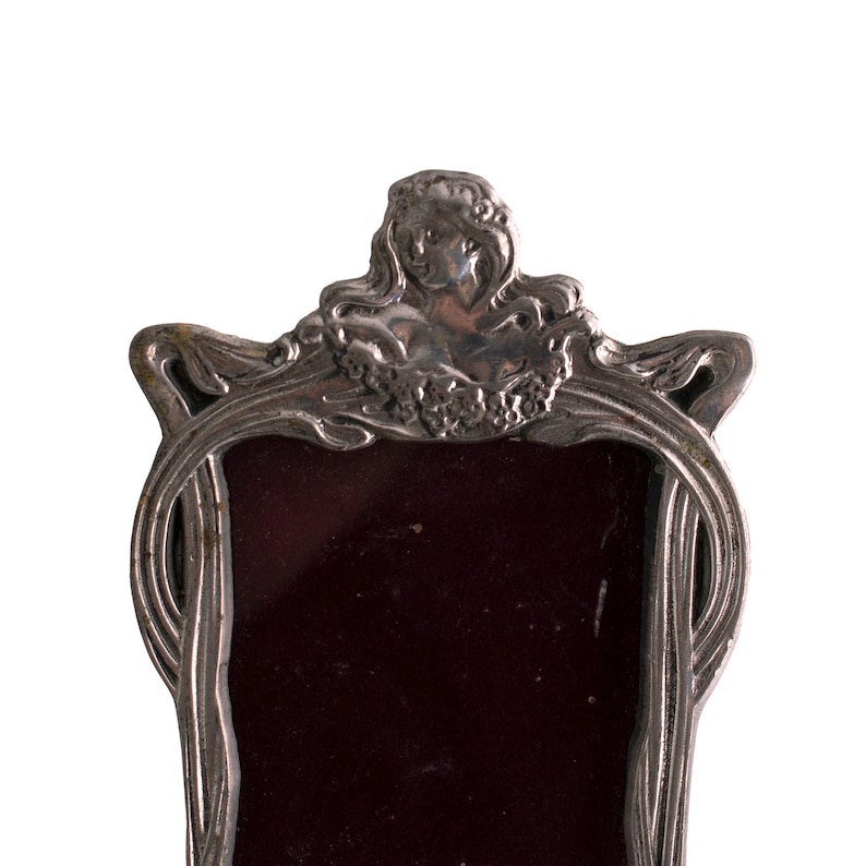 May include: A silver picture frame with an ornate design featuring a woman's face and floral motifs. The frame has a dark red velvet backing.