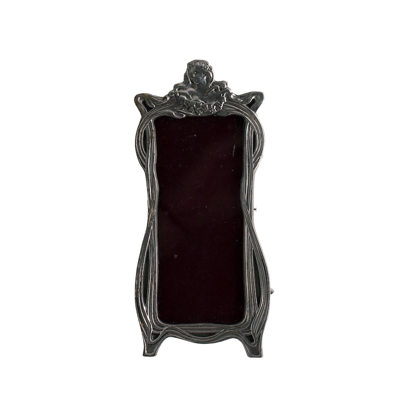 May include: A silver-toned picture frame with an ornate design featuring a stylized figure and floral motifs. The frame has a rectangular opening for a photo.