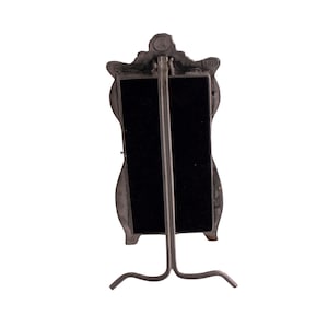 May include: A silver-toned metal mirror with a decorative frame in the shape of a woman's face. The mirror has a black backing and a stand.