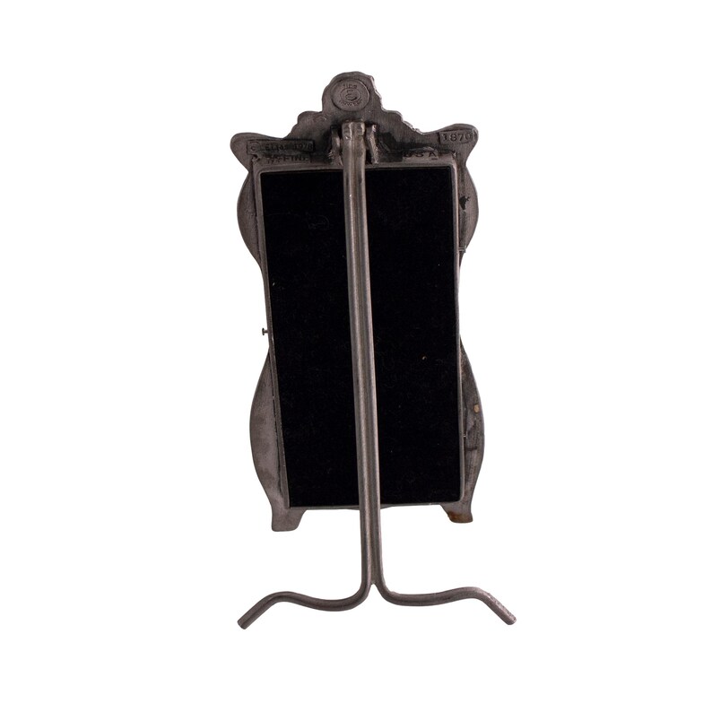 May include: A silver-toned metal mirror with a decorative frame in the shape of a woman's face. The mirror has a black backing and a stand.
