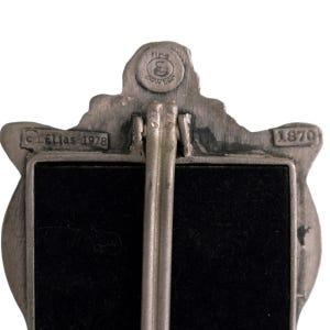 May include: A silver-toned metal picture frame with a black velvet backing. The frame features a detailed design of a lion's head with the text "Fine Pewter" and "1870" on the back. The frame is marked with "C Elias 1978".