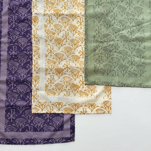 May include: An assortment of decorative fabric napkins in various colors. The napkins feature repeating patterns of stylized floral and religious symbols. Colors include purple, gold, and green. The napkins are likely made of linen or a similar fabric.