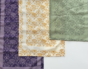 May include: An assortment of decorative fabric napkins in various colors. The napkins feature repeating patterns of stylized floral and religious symbols. Colors include purple, gold, and green. The napkins are likely made of linen or a similar fabric.
