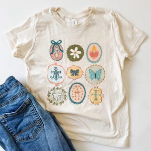 Puede incluir: Camiseta color crema con nueve ilustraciones ovaladas y coloridas. Las ilustraciones incluyen una flor, una mariposa, una cruz y un lazo. La camiseta se combina con vaqueros azules. La camiseta es una prenda informal.