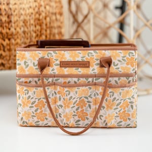 May include: A rectangular fabric storage caddy featuring a yellow and sage green floral pattern. The front includes three exterior pockets and a central rectangular patch with the text COME HOLY SPIRIT. It has two brown fabric handles.