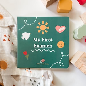 May include: A green children's book titled "My First Examen" with illustrations of a sun, heart, bird, and flower. The book is surrounded by wooden blocks and a patterned cloth.
