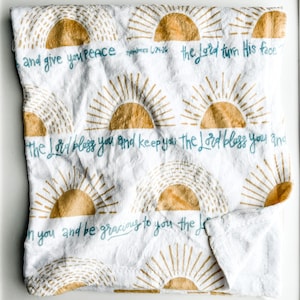 May include: White throw blanket with a repeating pattern of gold suns and the text "and give you peace numbers 6:24-26 the Lord turn His face the Lord bless you and keep you the Lord bless you and in you and be gracious to you the Lord".