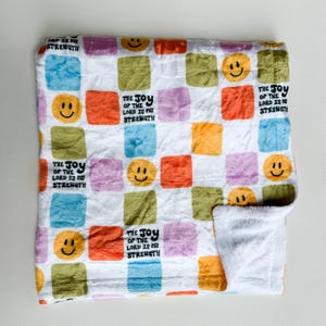 May include: A white fleece blanket with a colorful checkered pattern. The squares are in shades of yellow, orange, green, blue, and purple. Each square features a smiley face or the text "THE JOY OF THE LORD IS MY STRENGTH".