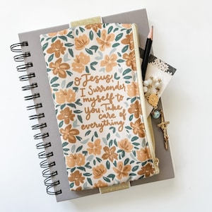 May include: A floral patterned zippered pouch with the text "O Jesus, I surrender myself to You. Take care of everything". The pouch is laying on top of a gray spiral bound notebook.