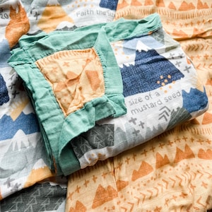 May include: A folded baby blanket with a green border and a center of orange fabric. The blanket features a mountain and arrow design in blue, gray, and orange, with the text "Faith the size" and "size of a mustard seed."