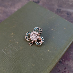 May include: A gold-tone shamrock brooch with green and clear rhinestones. The brooch is on a green book cover.