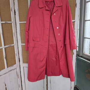 May include: A long-sleeved, pink, button-up coat with a collar. The coat has a single pocket on the left side and is hanging on a wooden hanger.