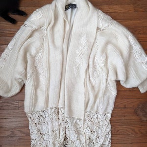 May include: A white knit cardigan with a long, flowing design. The cardigan features delicate floral embroidery and a lace trim.