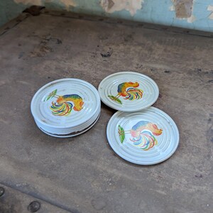 May include: Set of four white ceramic coasters with a colorful rooster design. The coasters are round and have a raised edge.