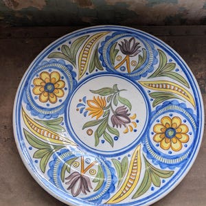 May include: A round, hand-painted ceramic plate with a floral design. The plate features a white base with a blue border and intricate patterns in yellow, green, and purple. The central design is a bouquet of flowers.