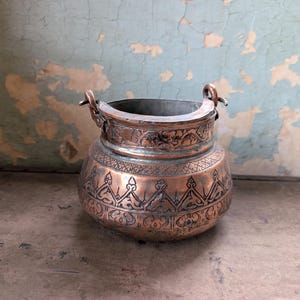 May include: A round copper pot with a wide opening and two small metal handles attached to the rim. The exterior surface features intricate engraved patterns, including geometric lines and stylized floral motifs, with a warm metallic finish.