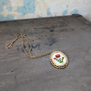 May include: Gold-toned necklace with an oval pendant featuring a hand-stitched red flower on a white background. The pendant is framed by a decorative gold border. The chain is delicate and the necklace is displayed on a dark surface.