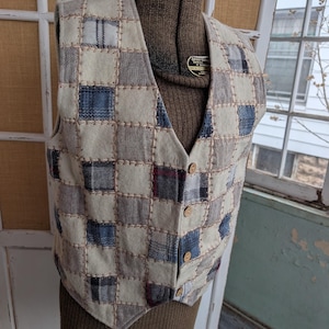 May include: A patchwork vest with a V-neck and wooden buttons. The vest features a pattern of squares in shades of cream, blue, gray, and burgundy, stitched together with tan thread. The vest is displayed on a mannequin.