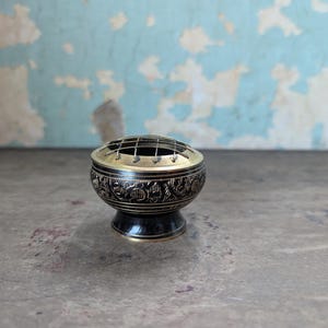 May include: A decorative incense burner with a black base and a gold-colored rim. The body of the burner is black with an embossed floral design. The top has a gold-colored rim with a wire grid.