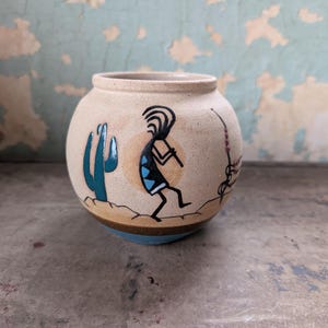 May include: A round, beige ceramic pot featuring a hand-painted black Kokopelli figure playing a flute, a teal saguaro cactus, and a thin brown line representing desert terrain. The base of the vessel is accented with a dark brown and blue band.