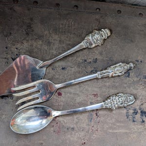 May include: Three antique silver serving utensils: a cake server, a fork, and a spoon. The handles are intricately detailed with ornate designs, and the blades and tines are polished to a high shine. The utensils are arranged on a textured, dark surface.