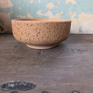 May include: A small, round, light tan ceramic bowl with a rough, porous, and textured exterior surface. The bowl features a simple, wide opening and a slightly raised circular base, showcasing a natural, unglazed, and earthy clay appearance.