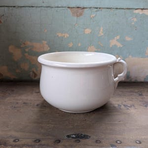 May include: A white, ceramic chamber pot with a rounded body and a small handle. The antique pot is set against a distressed, peeling blue wall and a weathered, metal-clad surface.