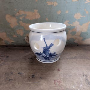 May include: A white ceramic onion keeper with a blue and white windmill design. The keeper has three circular holes and a blue trim. The background is a distressed, peeling wall.