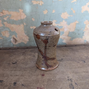 May include: A small, handmade ceramic vase with a speckled brown and tan glaze. The vase has a narrow neck, a flared base, and a single hole near the top. Dark brown markings decorate the surface. The vase is approximately 5 inches tall.