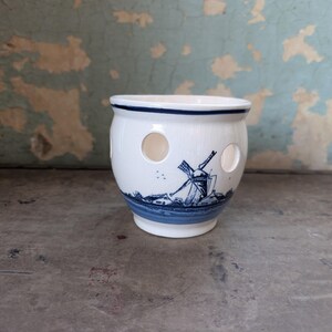 May include: A white ceramic onion pot with a blue windmill design and three circular holes. The pot has a blue band around the base and rim. The background is a distressed, peeling wall.