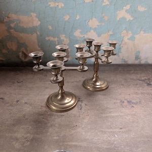 May include: Two brass candelabras, each featuring a central stem and four arms, totaling five candle holders per piece. The metal has a polished finish. A small oval metal plate at the bottom reads Belber Traveling Goods.