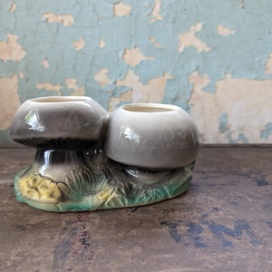 May include: A ceramic planter shaped like two mushrooms with a green and yellow base. The mushrooms are gray and have a smooth, glazed finish.