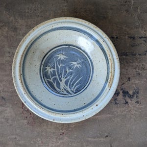 May include: A round, ceramic bowl with a light beige exterior and a blue interior. The bowl features a blue floral design in the center and two blue bands around the rim. The bowl is approximately 10 inches in diameter.