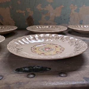 May include: A collection of vintage cream-colored plates with gold filigree and a floral center design. The plates have scalloped edges and are arranged on a weathered surface, suggesting a retro aesthetic.