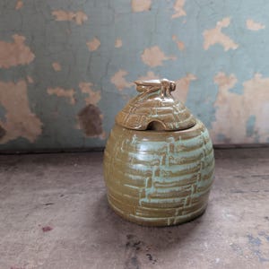 May include: A handmade ceramic jar with a lid, featuring a bee-shaped handle. The jar has a textured, beehive-like design with a blend of olive green and light blue glaze. The jar is approximately 5 inches tall.