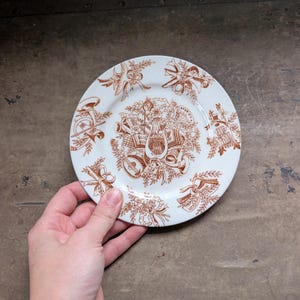 May include: A round white ceramic plate featuring a detailed brown transferware pattern. The center displays a cluster of musical instruments including a lyre, horn, and sheet music, surrounded by floral motifs and ribbons along the rim.