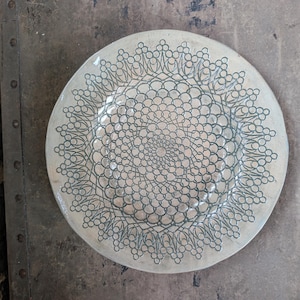 May include: A round, off-white ceramic plate with a detailed, symmetrical pattern in a darker gray color. The design features interconnected circles and floral-like shapes, creating a lace-like appearance. The plate is set against a textured, neutral-toned background.