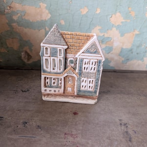 May include: A ceramic house figurine featuring a multi-gabled roof with tan shingles and a grey turret. The exterior walls are painted in muted blue and white with detailed window frames and a central tan door. The structure has a flat base.
