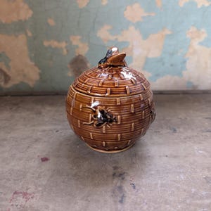 May include: A brown ceramic jar with a lid, featuring a woven basket design. The jar has a round shape and is adorned with small, dark-colored bee-like embellishments. The lid is topped with a small, stylized bee. The jar sits on a neutral surface.