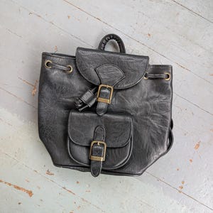 May include: Black faux leather backpack with two front pockets and gold-colored buckles. The backpack has a top handle and drawstring closure. The bag is sitting on a light blue and gray wooden surface.