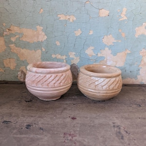 Puede incluir: Dos pequeñas macetas redondas de color beige con patrones tallados en forma de hojas alrededor de la parte superior. Las macetas están sobre una superficie oscura y desgastada, con una pared azul claro y beige descascarillada al fondo.