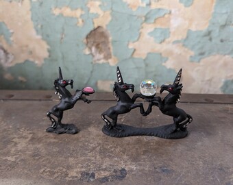 May include: Three black unicorn figurines, two of them facing each other and holding a clear crystal ball, the third unicorn is facing left and holding a pink heart. The figurines are on a dark brown surface.