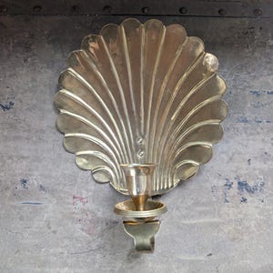 May include: A polished brass wall sconce featuring a large, fan-shaped scallop shell backplate with vertical ridges. A single candle cup is attached to the lower center of the shell, supported by a curved metal bracket extending from the base.