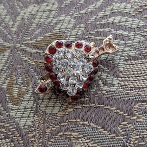 May include: A gold-tone brooch in the shape of a heart with an arrow through it. The heart is decorated with clear and red rhinestones.