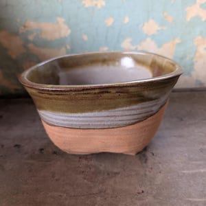 May include: A small, hand-thrown ceramic bowl with an irregular, organic shape. The upper portion features a glossy, olive-green and grey glaze, while the lower base remains unglazed, revealing the natural, textured terracotta-colored clay.