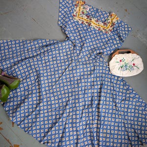 May include: A vintage blue dress with a white floral pattern, paired with green and purple shoes and a white purse with a wooden handle. The dress features a detailed neckline with floral embroidery, creating a cohesive ensemble.