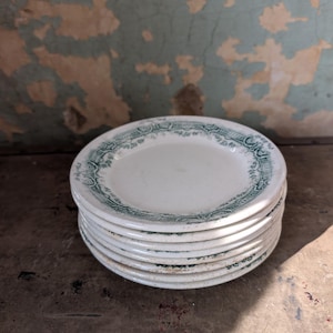 May include: A stack of white ceramic plates with a green floral border design. The plates are stacked on top of each other, showcasing the intricate details of the decorative edge. The plates are a vintage style.