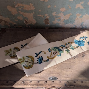 May include: Two off-white fabric panels with embroidered floral designs in shades of blue, green, and gold. The panels are rectangular and appear to be decorative elements, possibly for a pillow or other textile project. They are placed on a vintage trunk.
