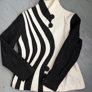 May include: A black and white jacket with a unique design. The jacket features a high collar and three black buttons. The left side has black and white wavy stripes, while the right side is solid white. The sleeves are solid black.